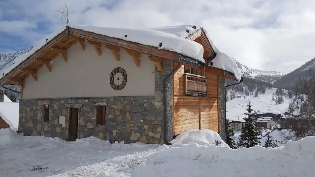 Façade en pierre du Chalet Kergwen à Montgenèvre pour 18 personnes sous la neige, vue de la rue avec parking et montagne