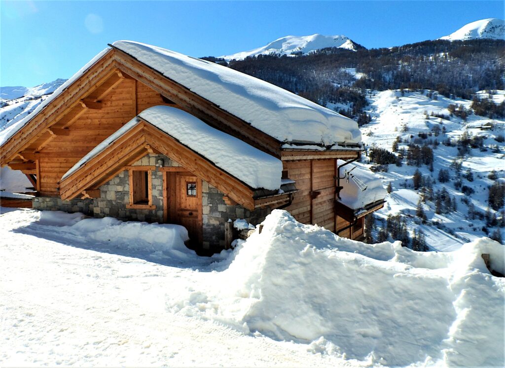 Façade principale en bois du chalet à Vars sous la neige avec le parking enneigé, les pistes de ski et les montagnes en arrière-plan en hiver.