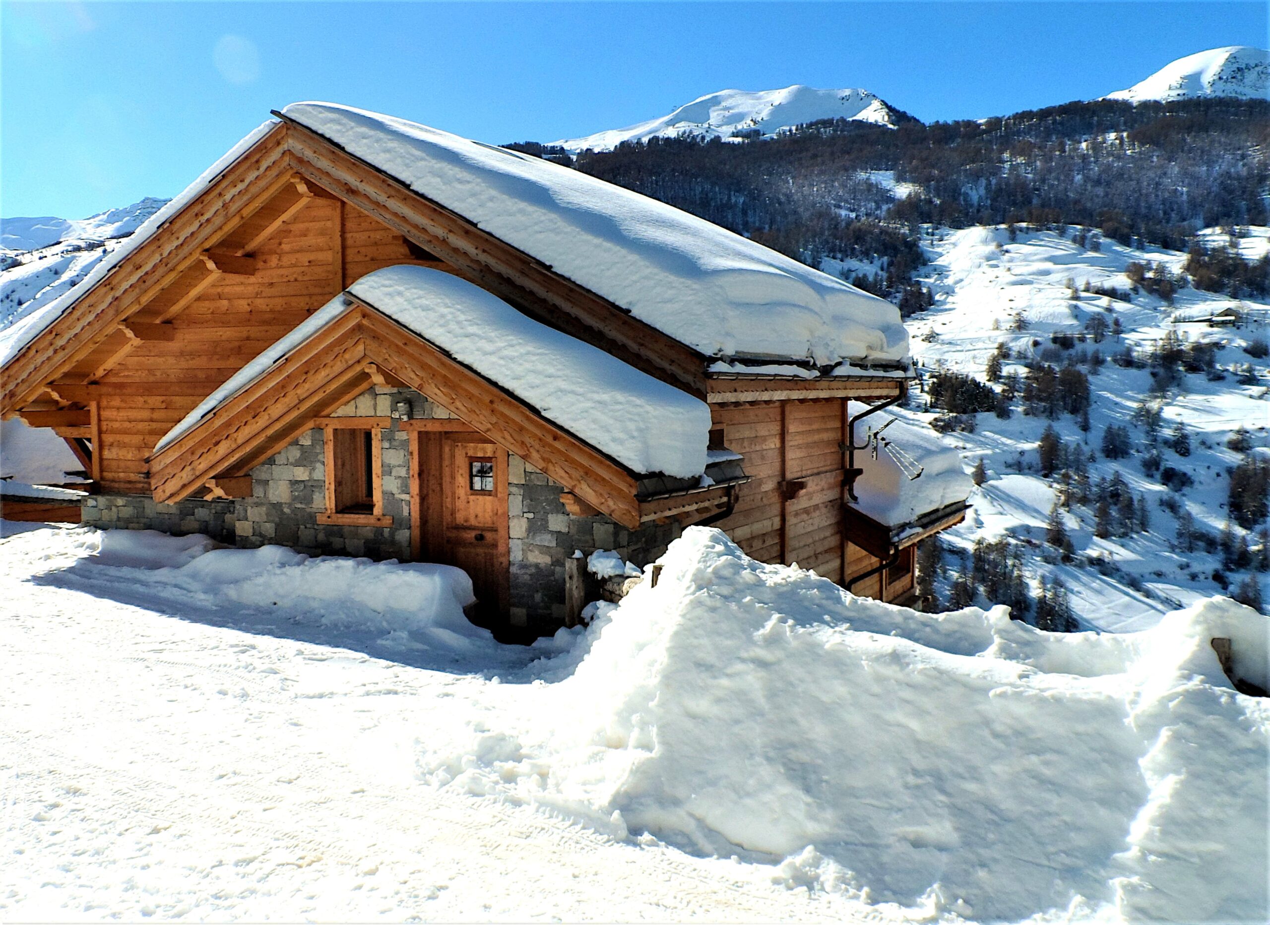 Façade principale en bois du chalet à Vars sous la neige avec le parking enneigé, les pistes de ski et les montagnes en arrière-plan en hiver.