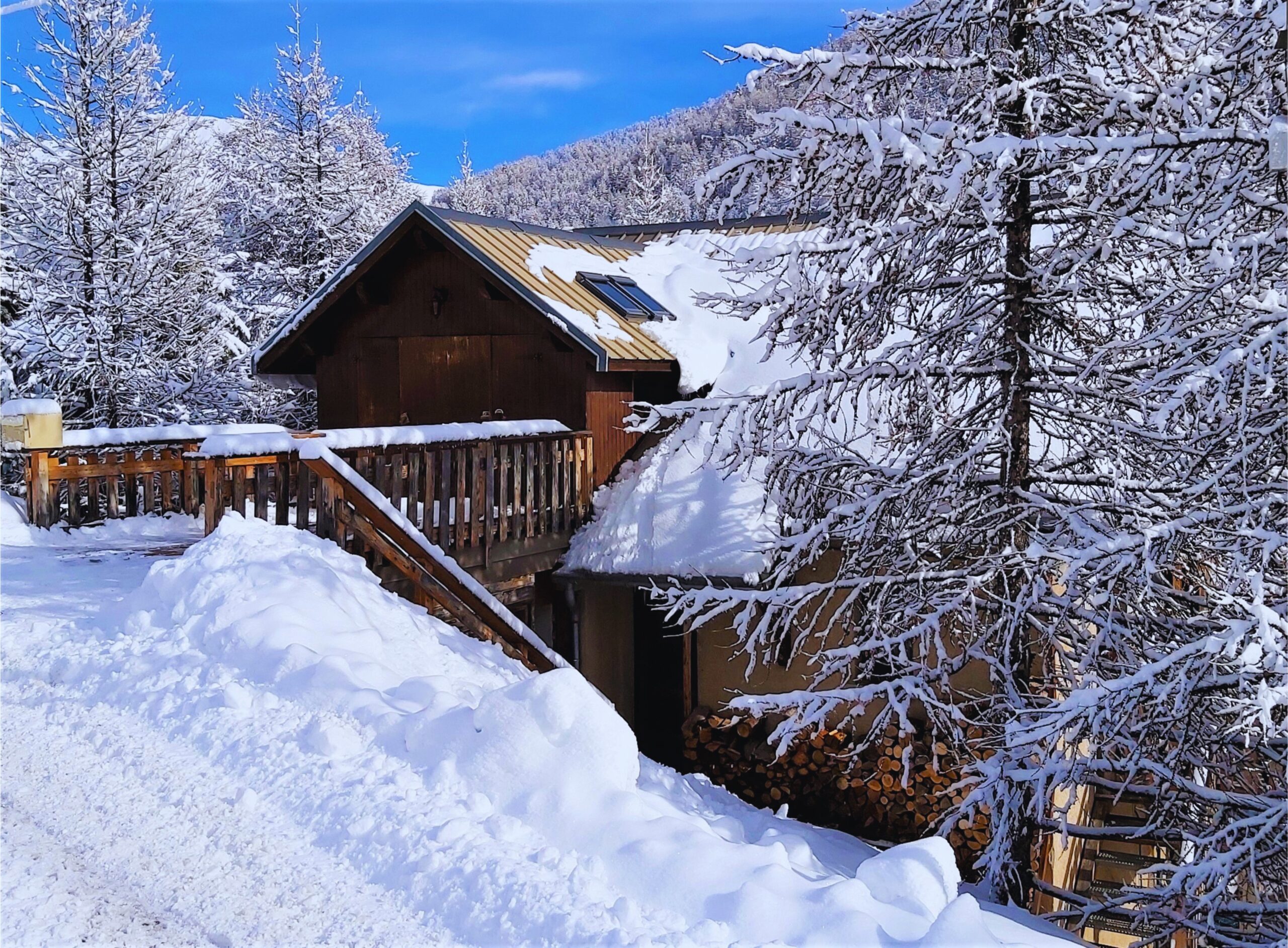 Vue du Chalet Le Mouflon à Vars depuis la rue en hiver, avec passerelle d’accès, zone de stationnement et bois stocké pour la cheminée.