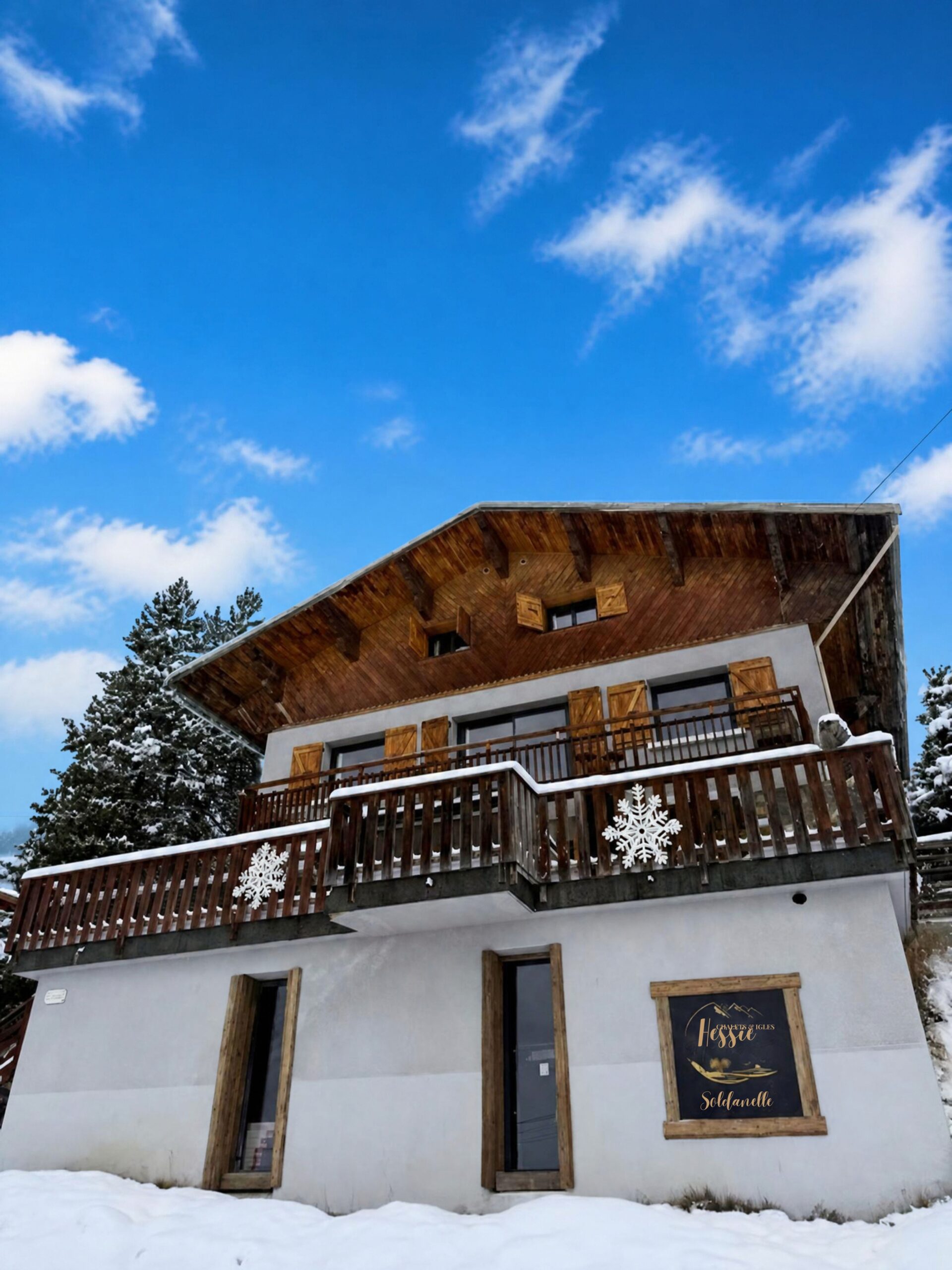 Façade du chalet La Soldanelle à Montgenèvre en hiver, avec balcons en bois, volets et sapins enneigés sous un ciel bleu