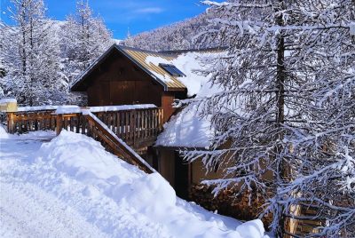 Vue du Chalet Le Mouflon à Vars depuis la rue en hiver, avec passerelle d’accès, zone de stationnement et bois stocké pour la cheminée.